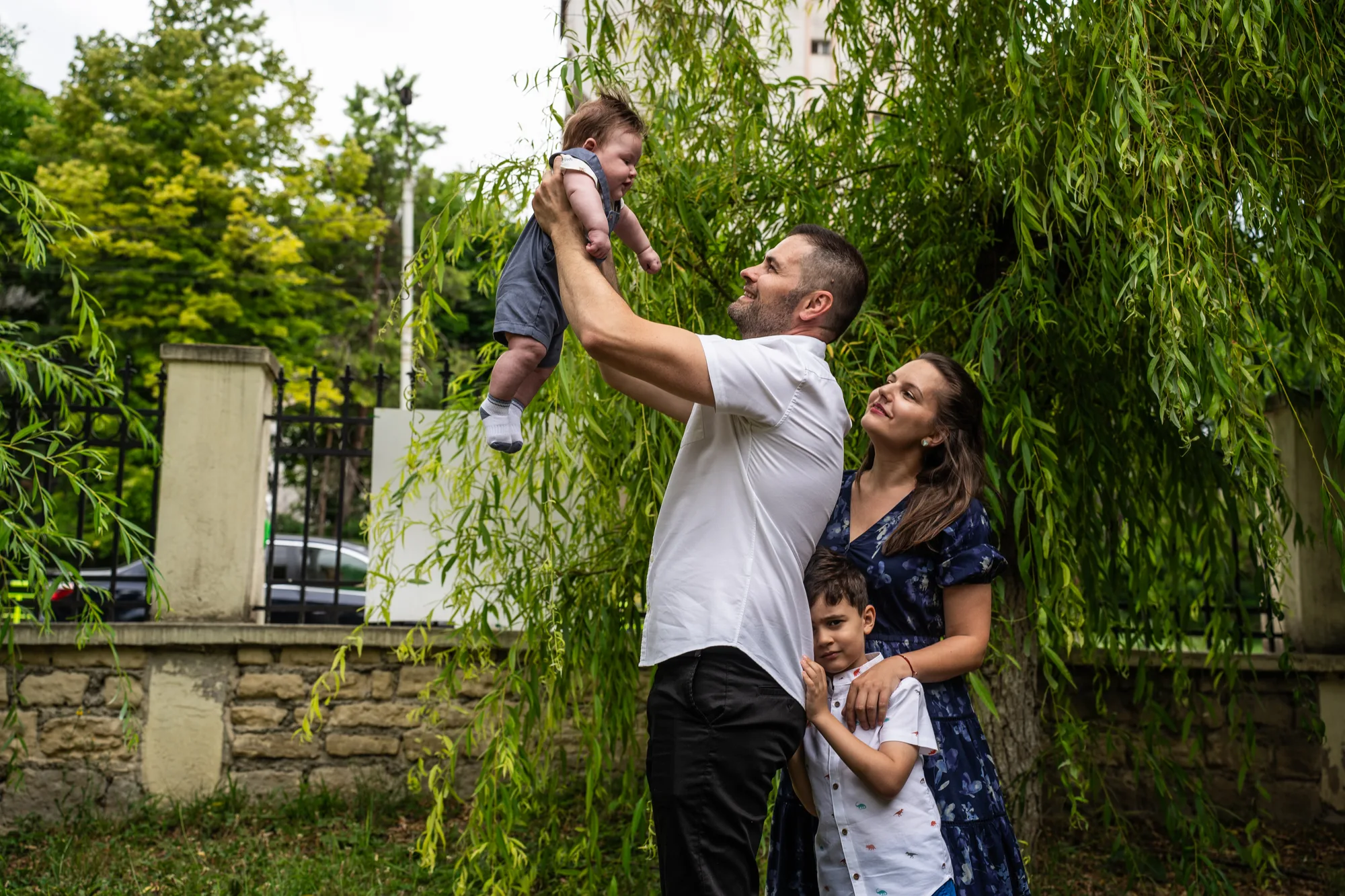 Bărbat ridicând un prunc deasupra capului, lângă o femeie și un băiat, într-o curte cu salcie