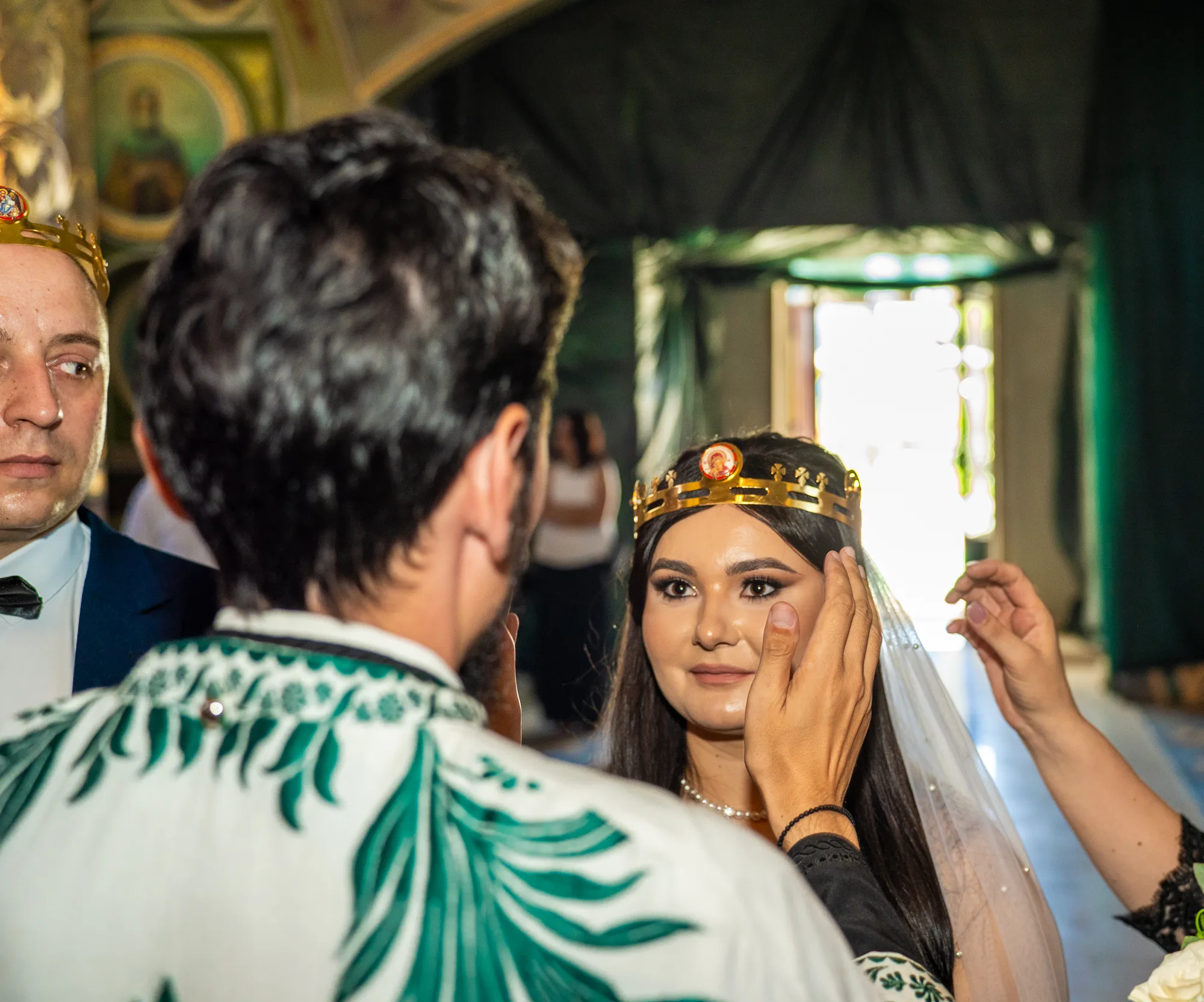 Mireasă cu cununie aurită pe cap, în biserică, cu o mână așezându-i vălul; lângă ea, un bărbat purtând cununie