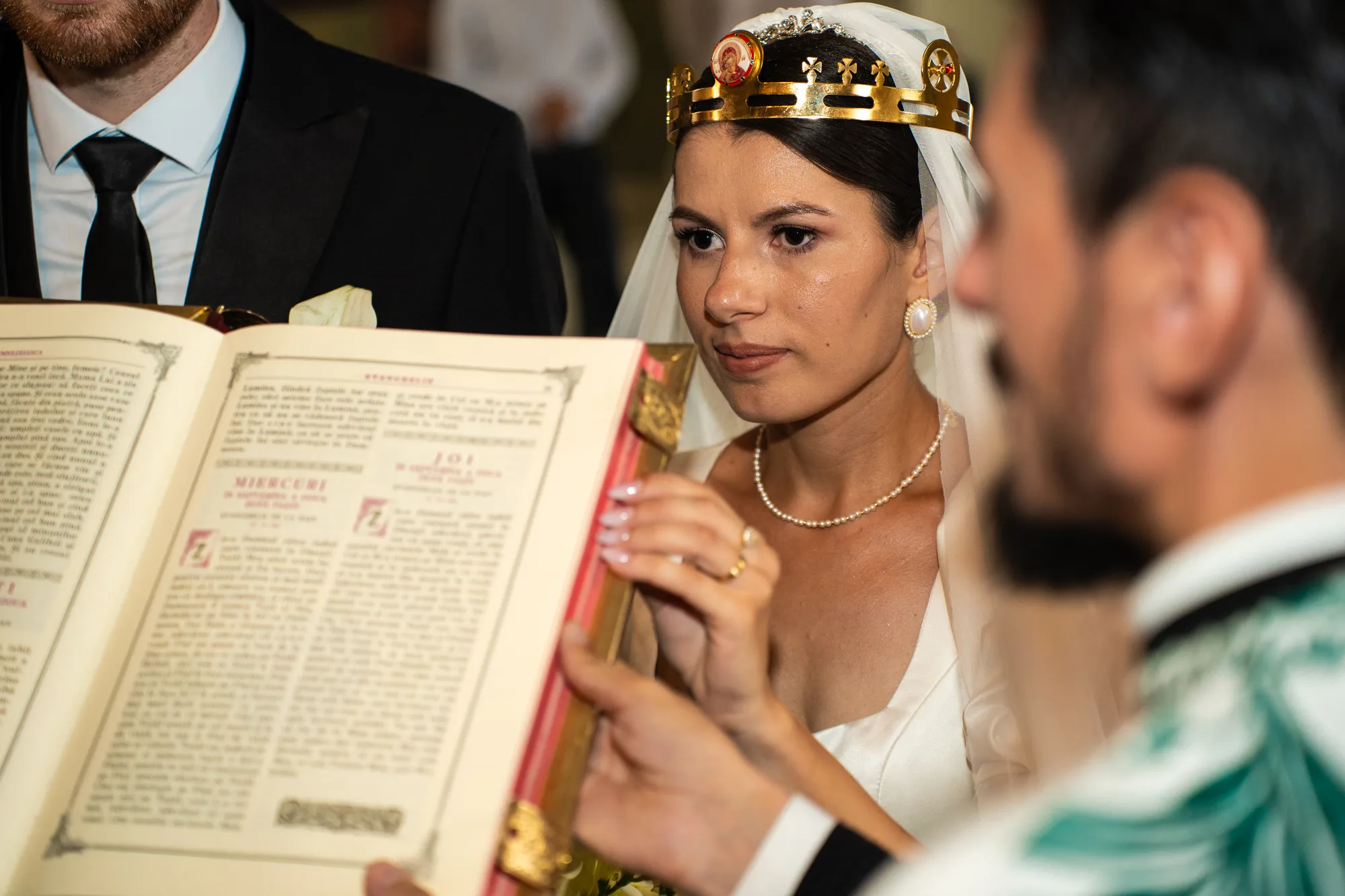 Mireasa cu cununia de aur pe cap, privind spre preotul care îi ține Evanghelia deschisă în față, cu mirele alături
